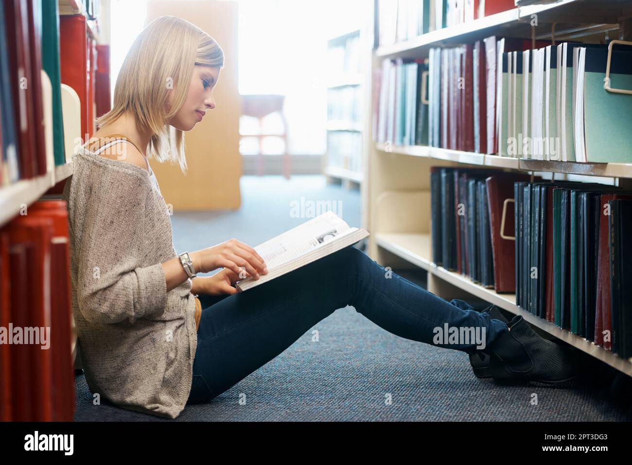 Studiando per il mio esame finale. Una giovane donna seduta sul pavimento e che legge in una biblioteca Foto Stock