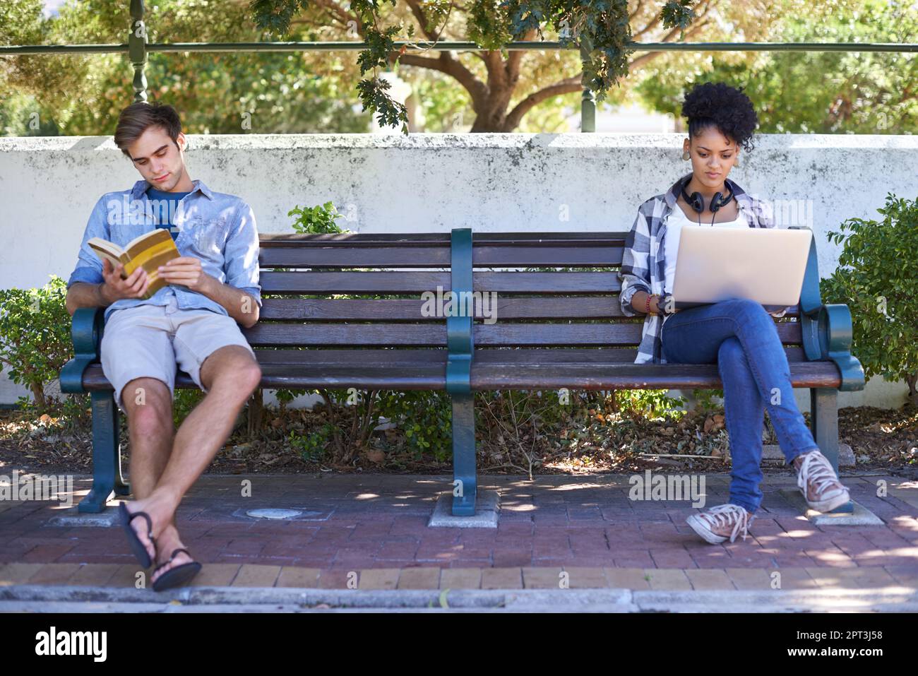 Perso nei nostri mondi separati. due studenti universitari che studiano mentre si siedono su una panchina del campus Foto Stock