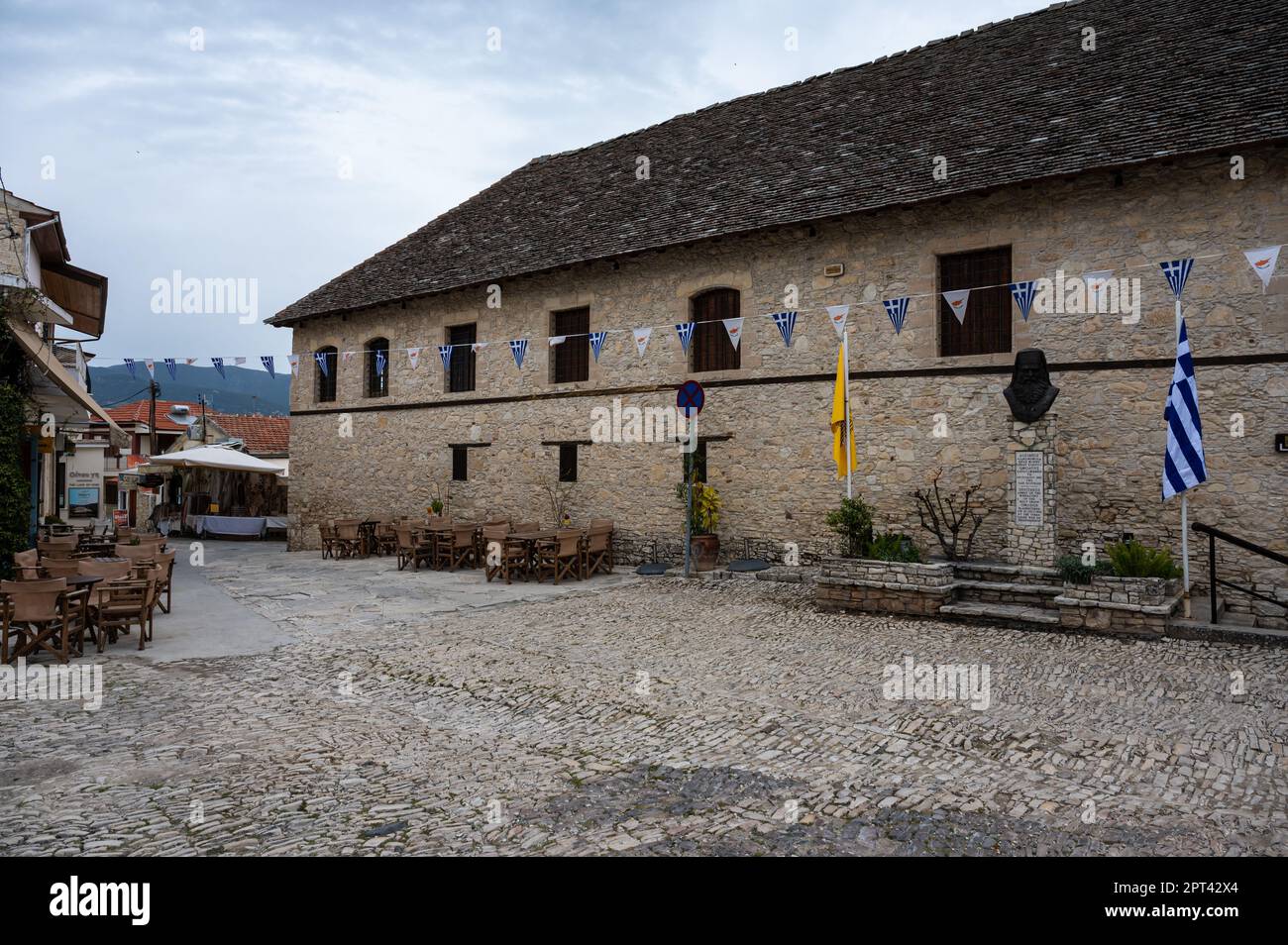 Omodos, Distretto di Limassol, Cipro, 24 marzo 2023 - tradizionale muro di villaggio con pavimento in pietra di ciottoli e bandiere cipriote e greche Foto Stock
