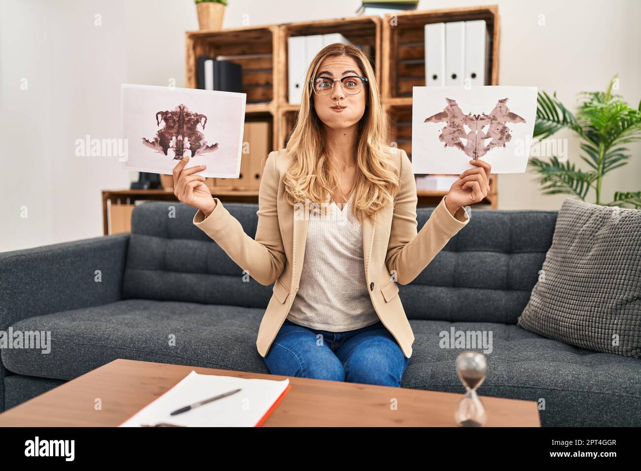 Giovane donna therapist bionda che tiene rorschach prova le guance puffing con faccia divertente. bocca gonfiata con aria, che cattura l'aria. Foto Stock