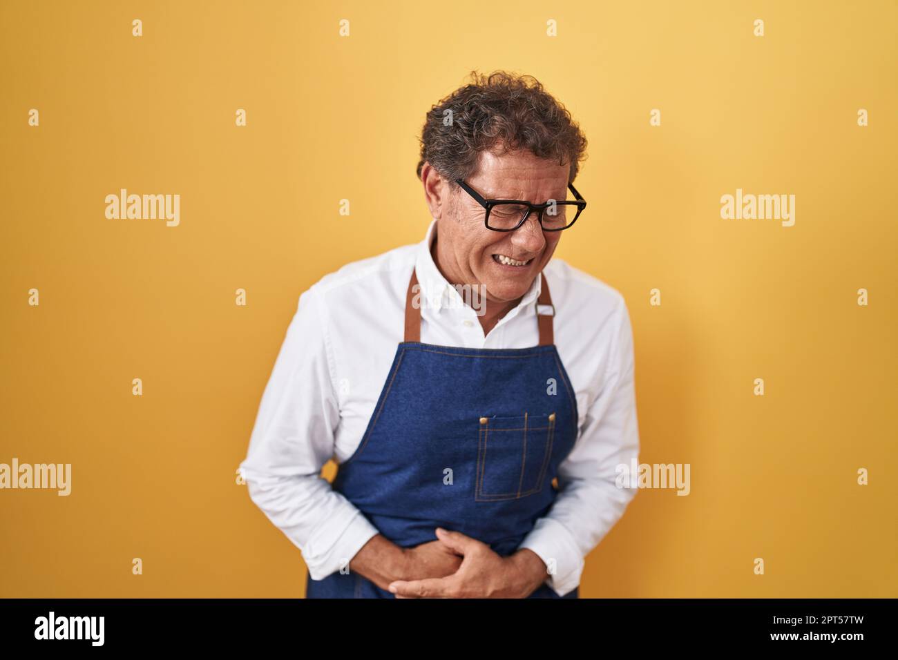 Uomo ispanico di mezza età che indossa un grembiule cuoco professionale con la mano sullo stomaco perché nausea, dolorosa sensazione di malessere. concetto di ache. Foto Stock