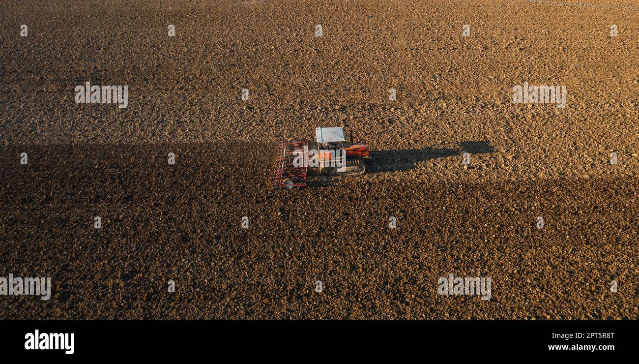 il contadino che guida un trattore cingolato trascina dietro una nuvola di polvere, arando, erpicando la rottura della crosta superficiale per facilitare la nascita e la semina di o Foto Stock