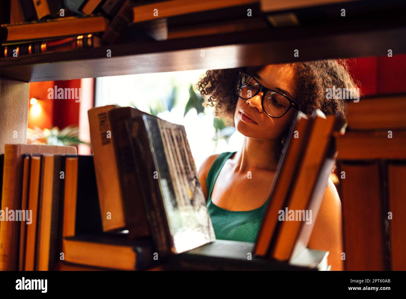 Felice studentessa afro-americana studia in biblioteca universitaria e cerca il libro, indossando gli occhiali Foto Stock