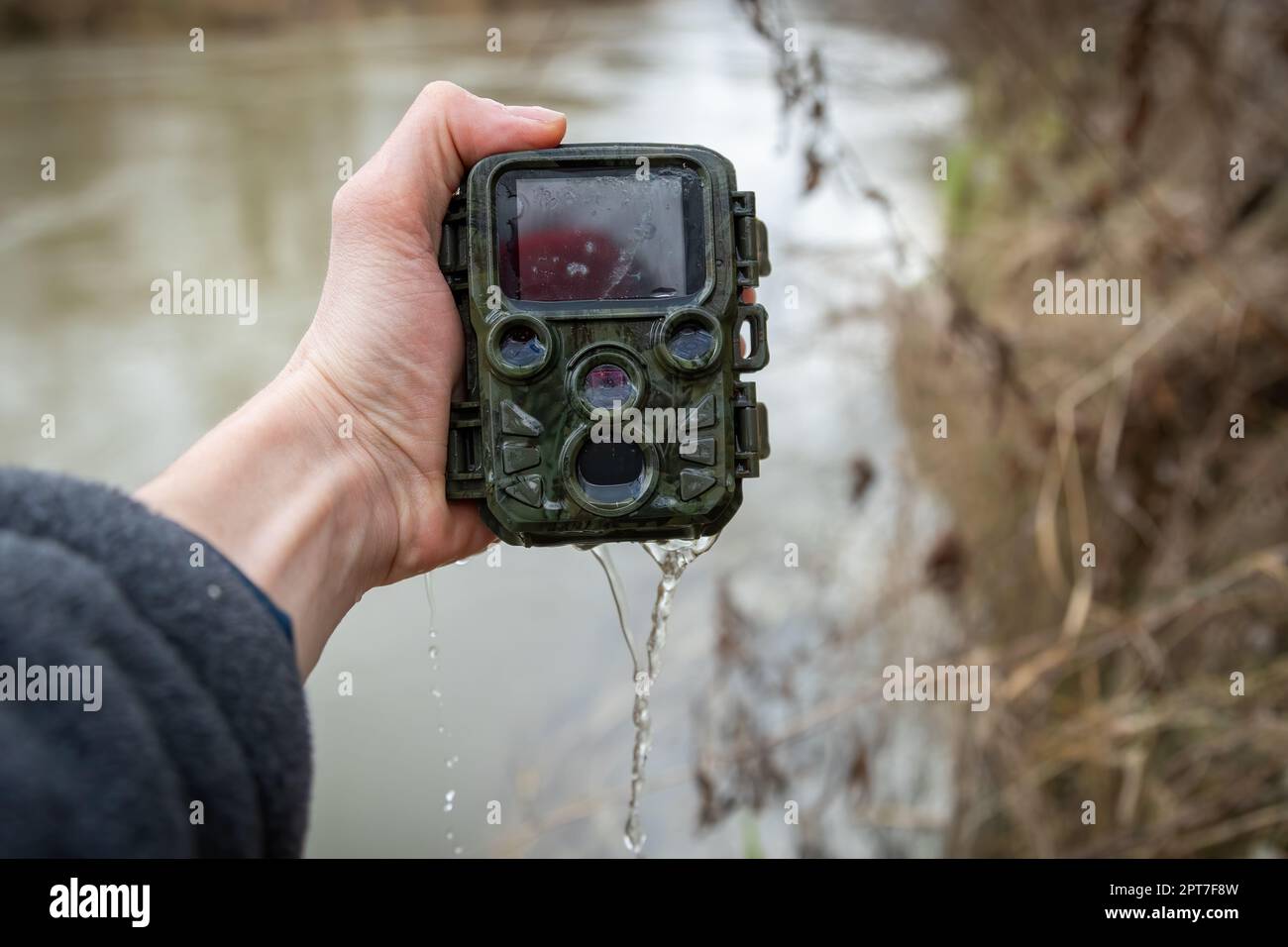 Mano che tiene una macchina fotografica del sentiero annegata vicino al  fiume con acqua che perde dentro. Trappola della fotocamera danneggiata con  sensore di movimento sulla riva del fiume. Dispositivo rotto per