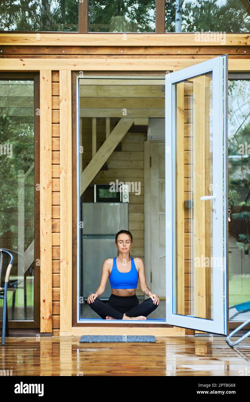 Giovane donna rilassata siede in posa lotus yoga all'interno della cabina di legno di fronte alla porta aperta Foto Stock