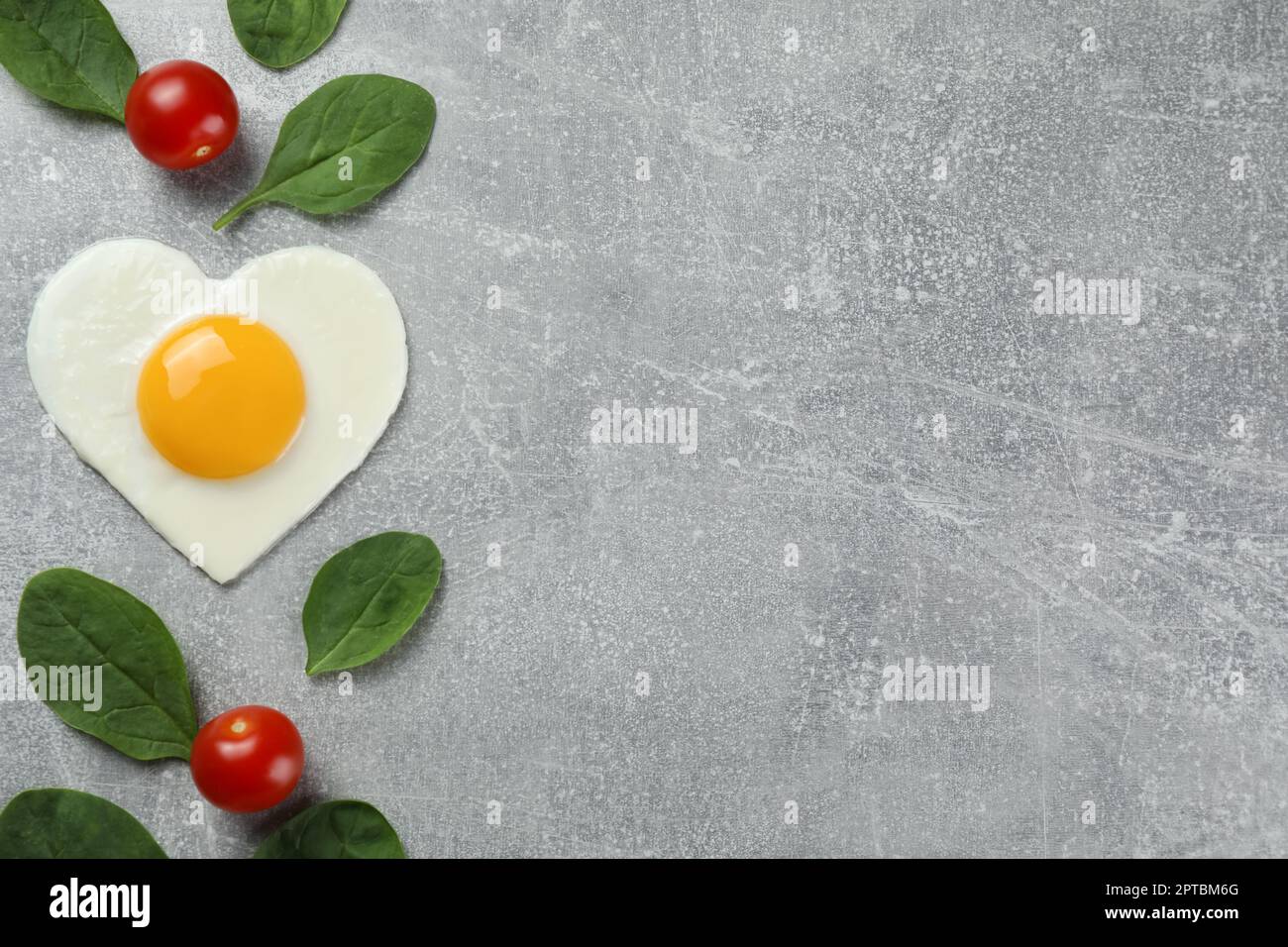 Uovo fritto a forma di cuore, pomodori e spinaci su tavolo grigio chiaro, piatto. Spazio per il testo Foto Stock