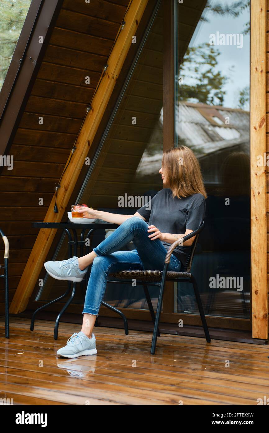 Donna si siede in una sedia di vimini vicino al tavolo sulla terrazza di legno della sua cabina forestale Foto Stock