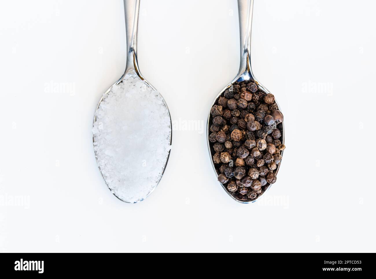 Un cucchiaio pieno di sale da tavola e grani di pepe nero su sfondo bianco Foto Stock