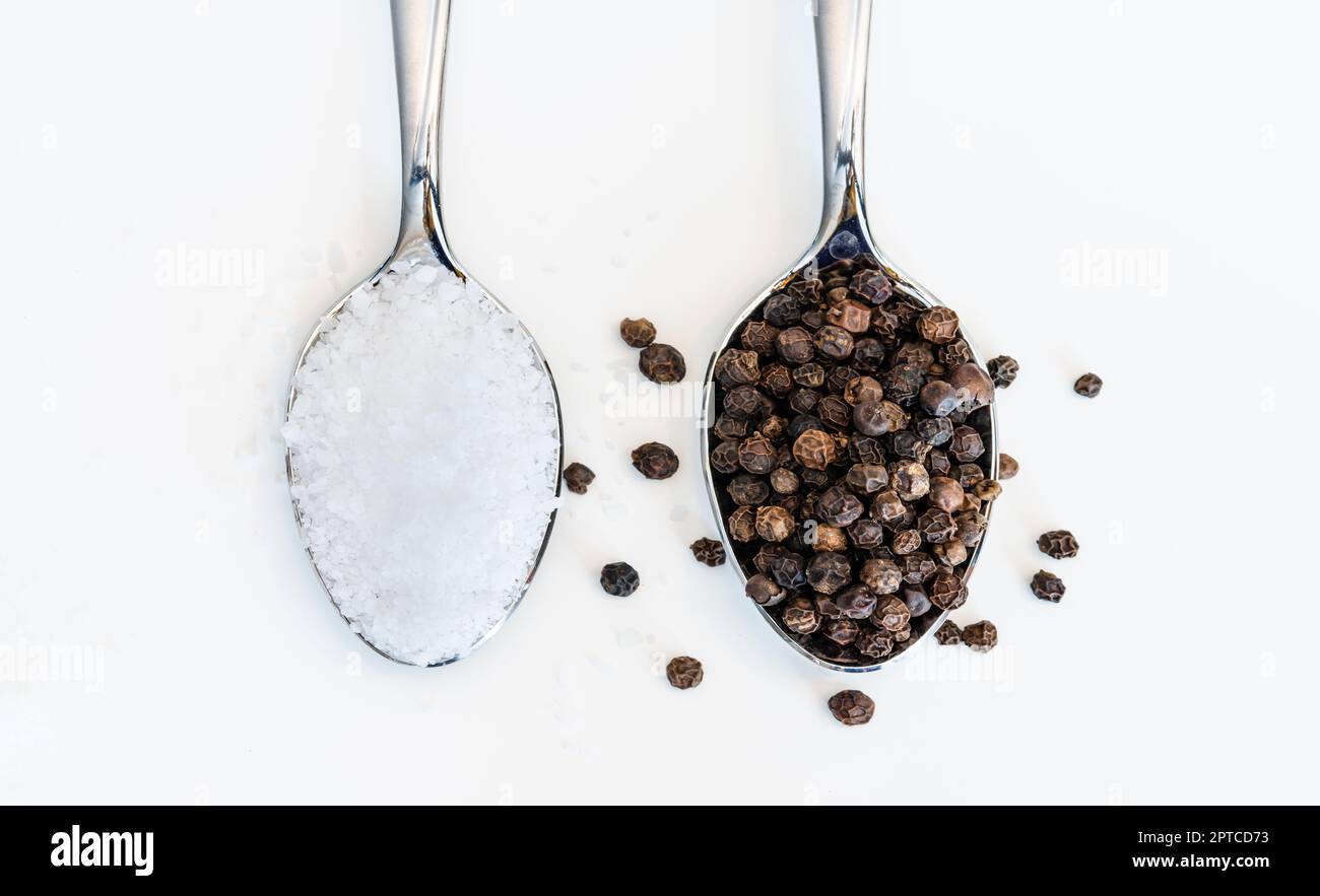 Cucchiaio traboccante pieno di sale da tavola e grani di pepe nero su sfondo bianco Foto Stock