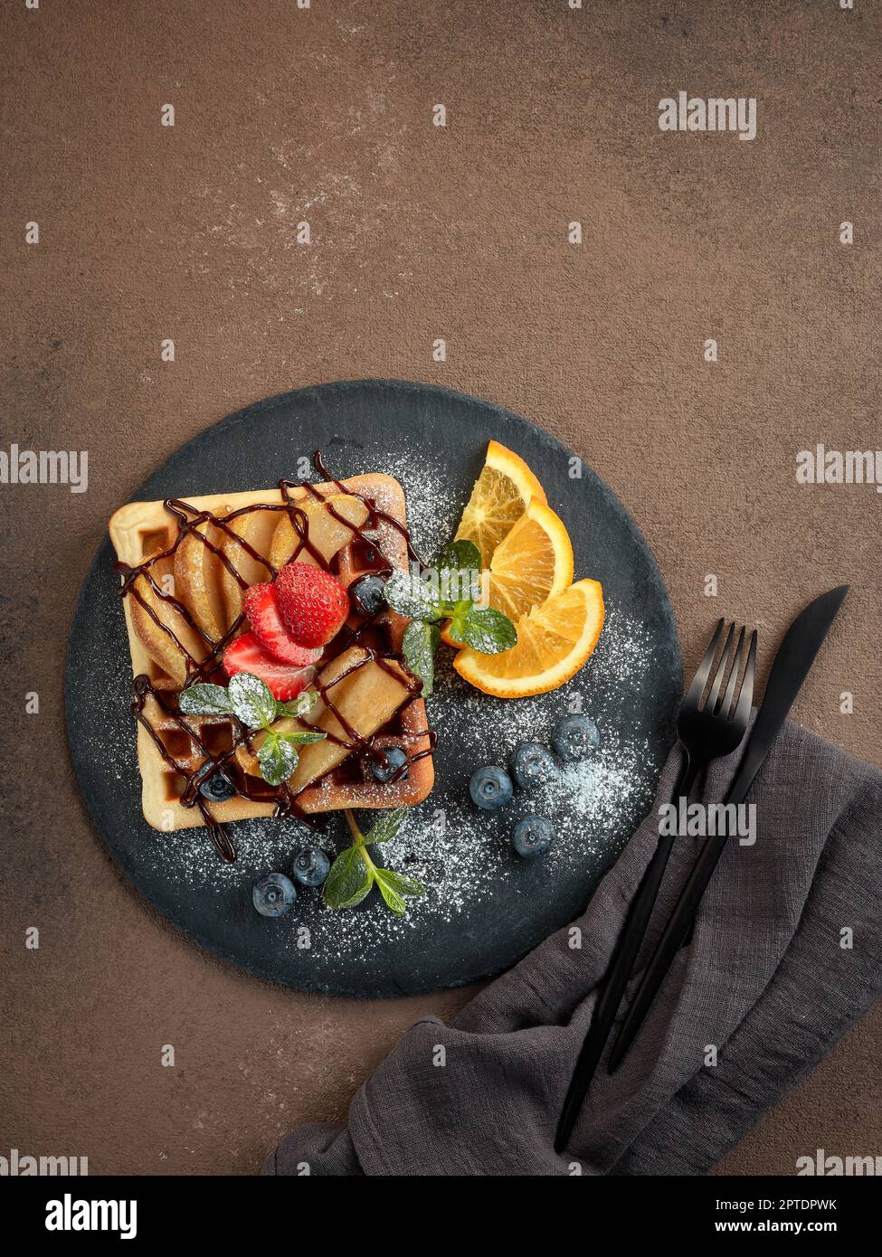 Deliziosi waffle con pera caramellata, frutti di bosco, menta, zucchero a velo e salsa dolce sul piano di servizio nero su sfondo marrone scuro. Dessert. Servizio Foto Stock