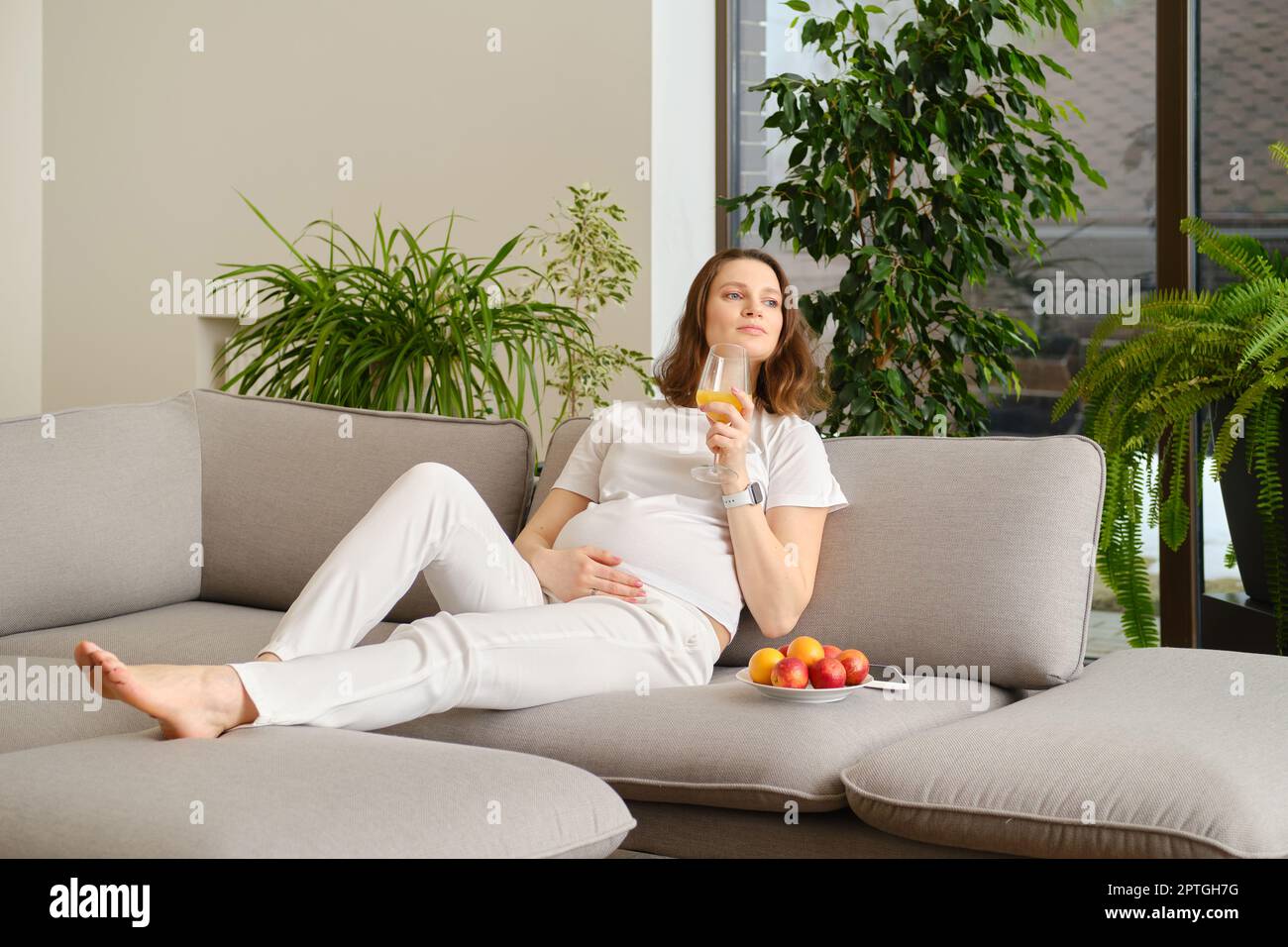 Donna incinta carina che riposa seduto sul divano a casa con un bicchiere di succo d'arancia fresco in mano Foto Stock