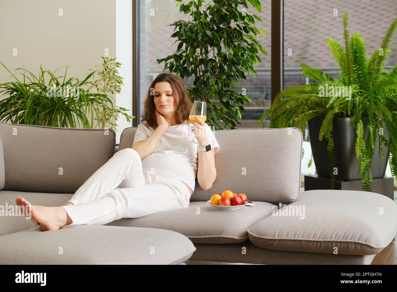 Donna incinta penosa che si riposa seduto sul divano a casa con un bicchiere di succo d'arancia fresco in mano Foto Stock