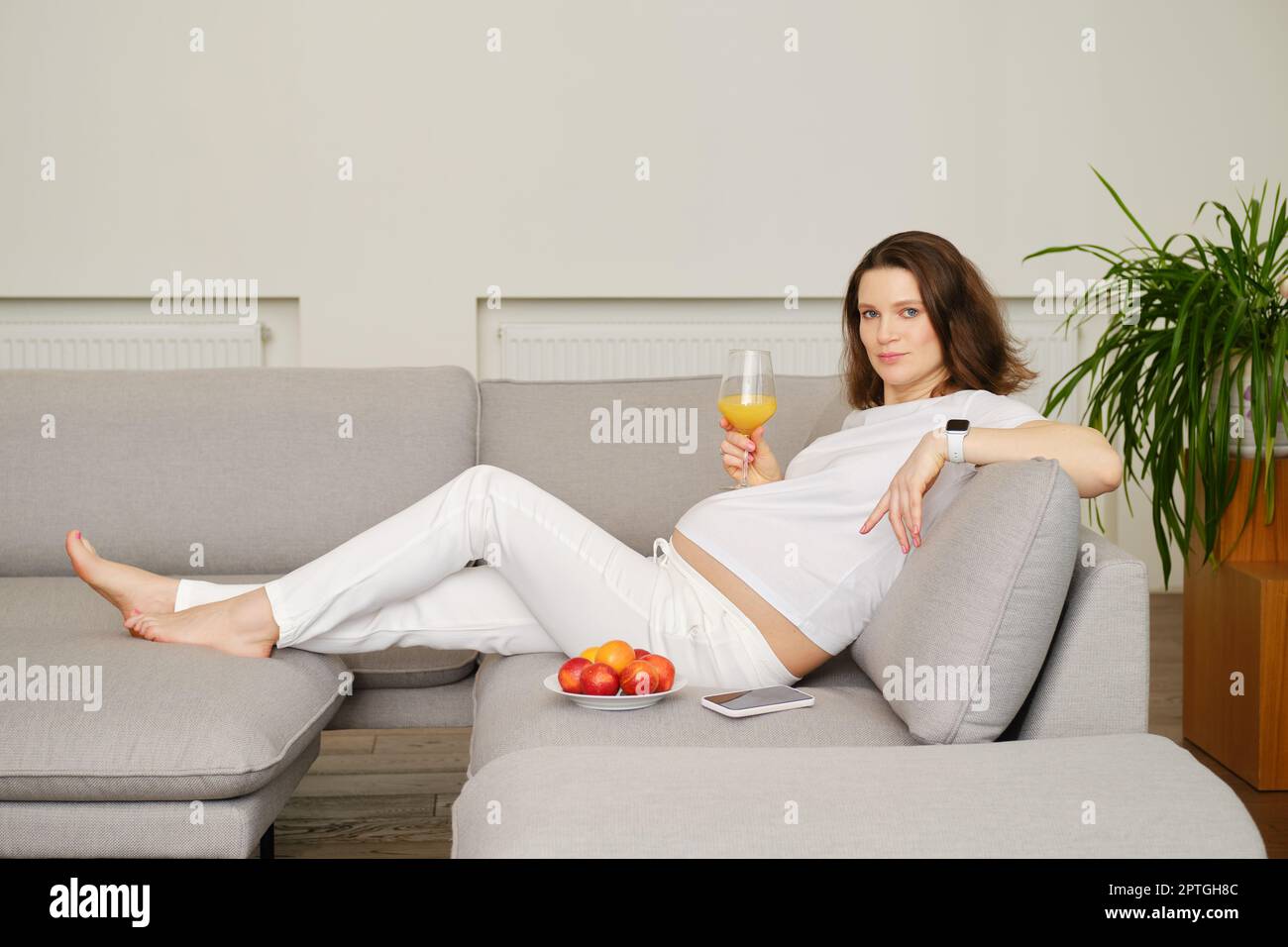 Donna incinta positiva sdraiata sul divano a casa con un bicchiere di succo d'arancia fresco in mano. Telefono cellulare e piatto con frutta accanto a lei. Foto Stock