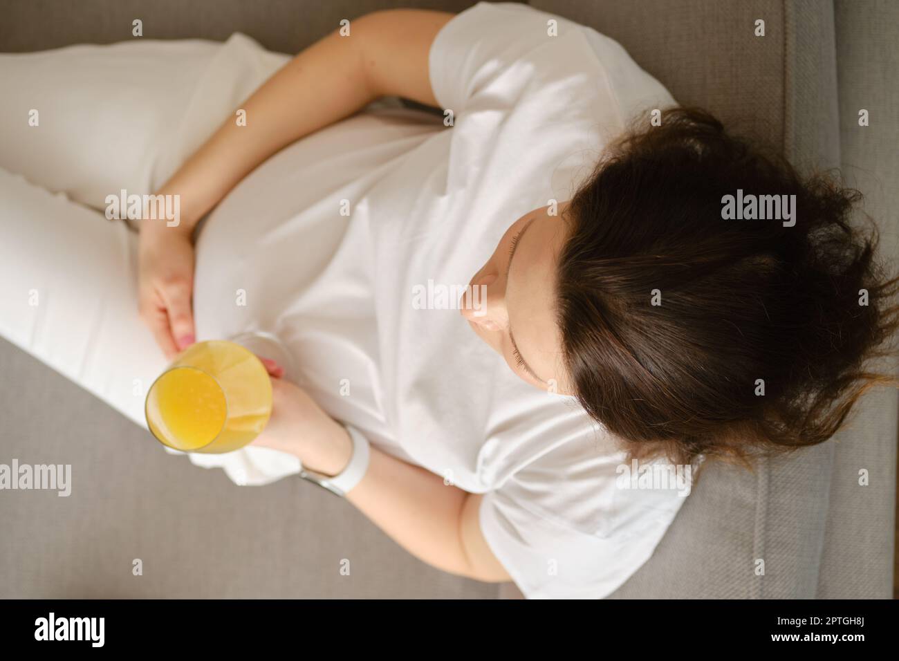 Vista dall'alto di una donna incinta seduta sul divano con un bicchiere di succo d'arancia fresco in mano Foto Stock