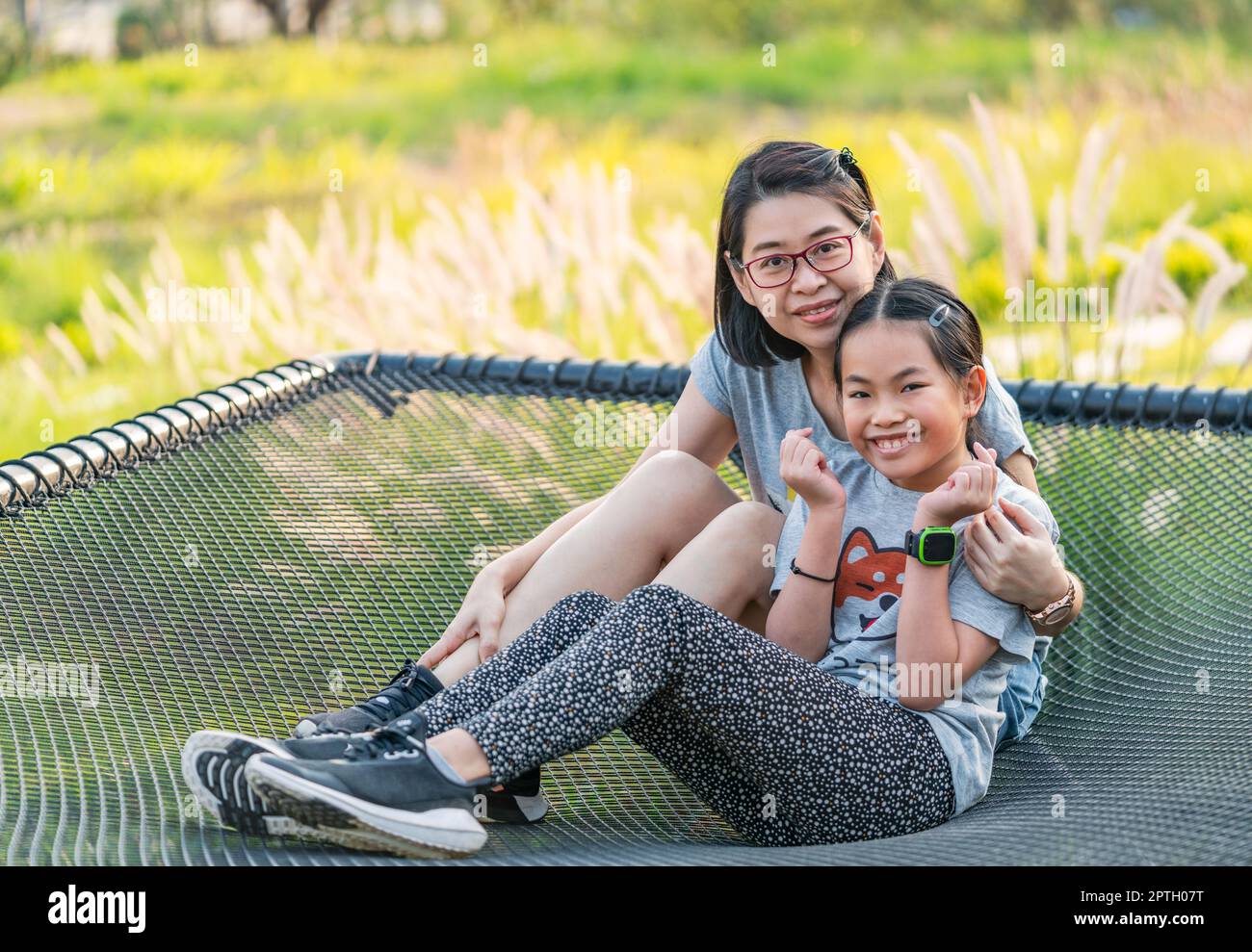 Ritratto di madre single asiatica e piccola figlia seduto su una grande rete di seduta in un resort, sfondo sfocato di fiori d'erba, luce naturale, sorridente fac Foto Stock
