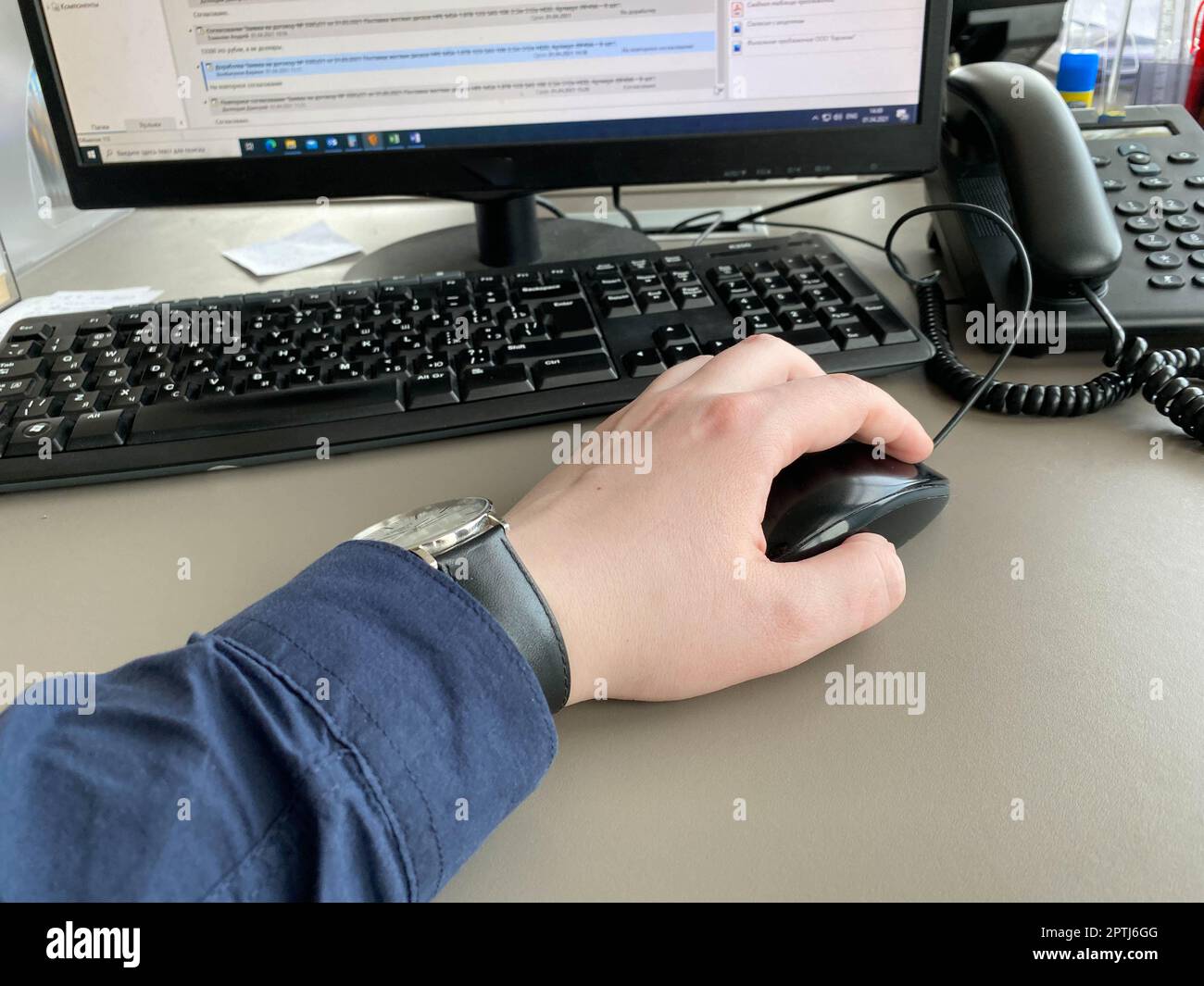 Una mano dell'uomo con una camicia e un orologio tiene un mouse del calcolatore sul tavolo al tavolo dell'ufficio con un calcolatore con una tastiera. Lavoro aziendale. Foto Stock