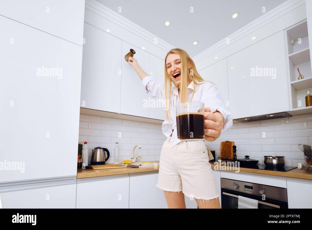 Donna allegra che balla e urla con una tazza di caffè in mano in cucina Foto Stock