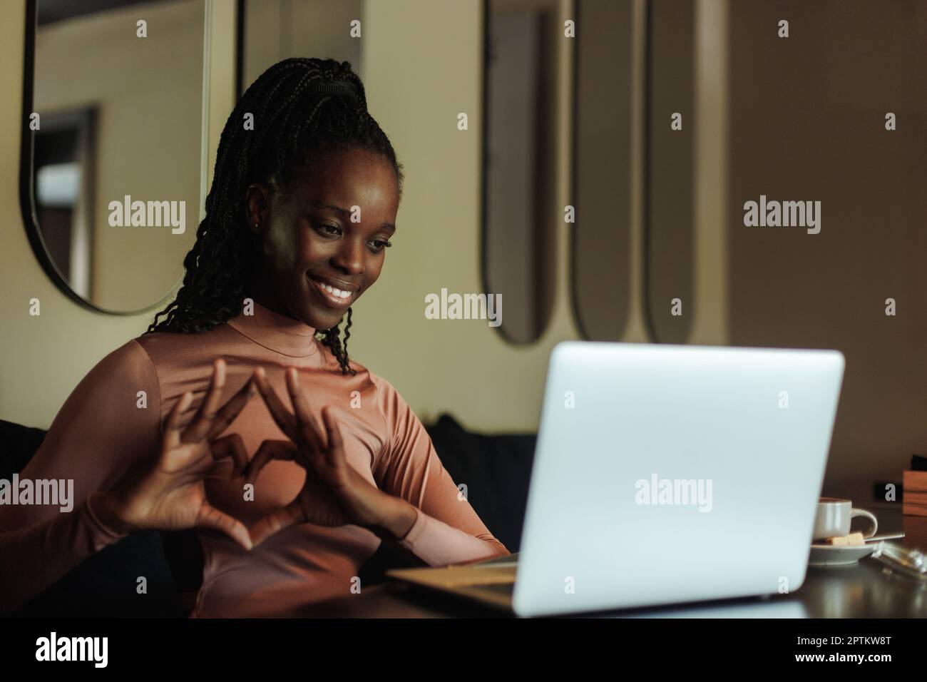 Ritratto di una giovane donna afroamericana sorridente seduta al tavolo vicino al computer portatile, che mostra il cuore con le dita sullo schermo. Foto Stock