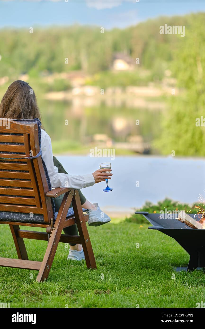 Giovane donna con un bicchiere di sangria in mano si rilassa in natura seduto accanto ad un barbecue, vista posteriore Foto Stock