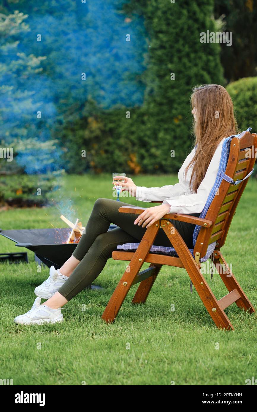 Giovane donna che riposa in poltrona sul cortile accanto a un barbecue, vista sul profilo Foto Stock