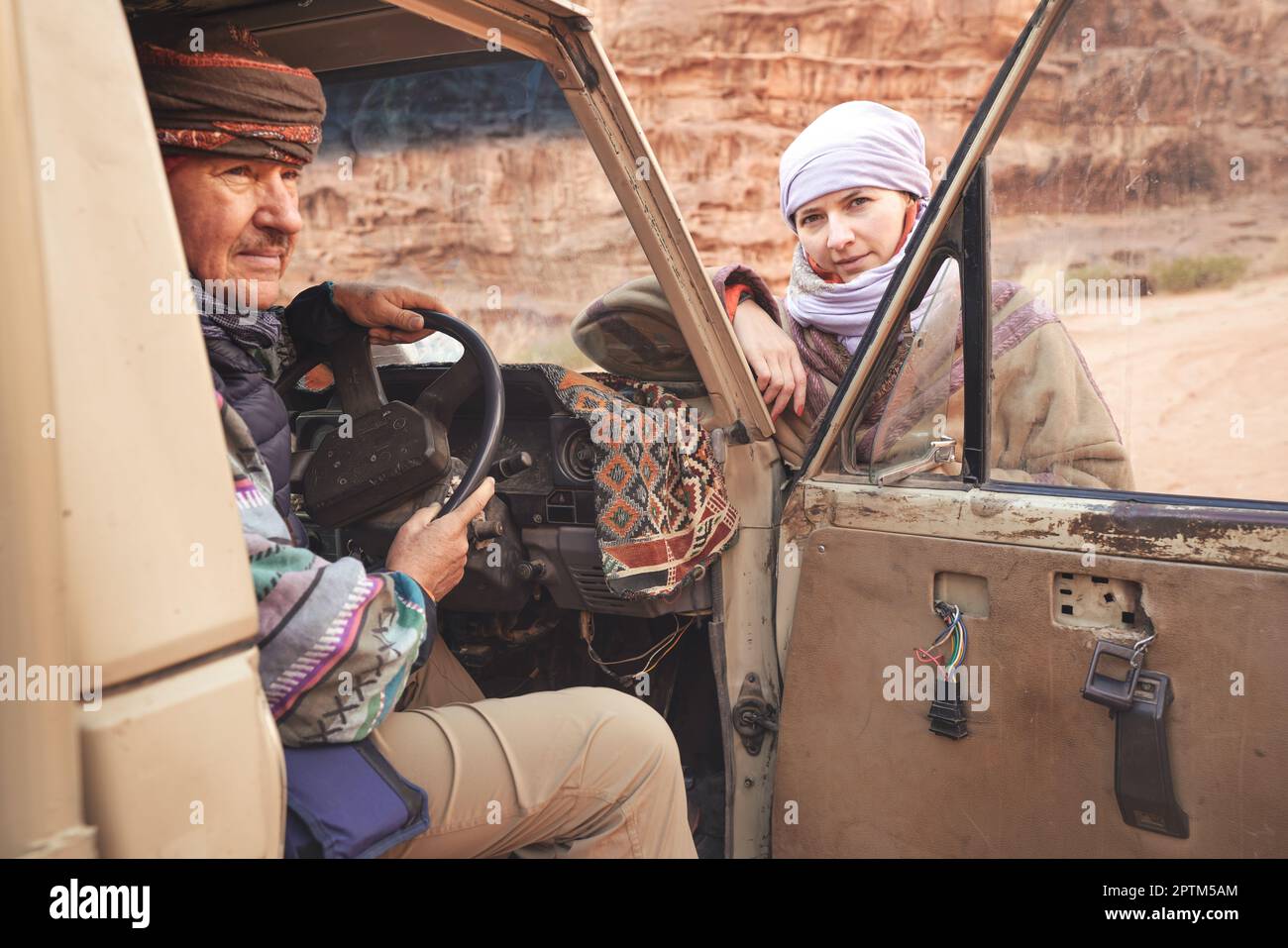 Uomo in cappotto beduino tradizionale - bisht - e foulard, posando dietro al volante in vecchio veicolo 4WD, guardando al lato, giovane donna e paesaggio desertico o Foto Stock