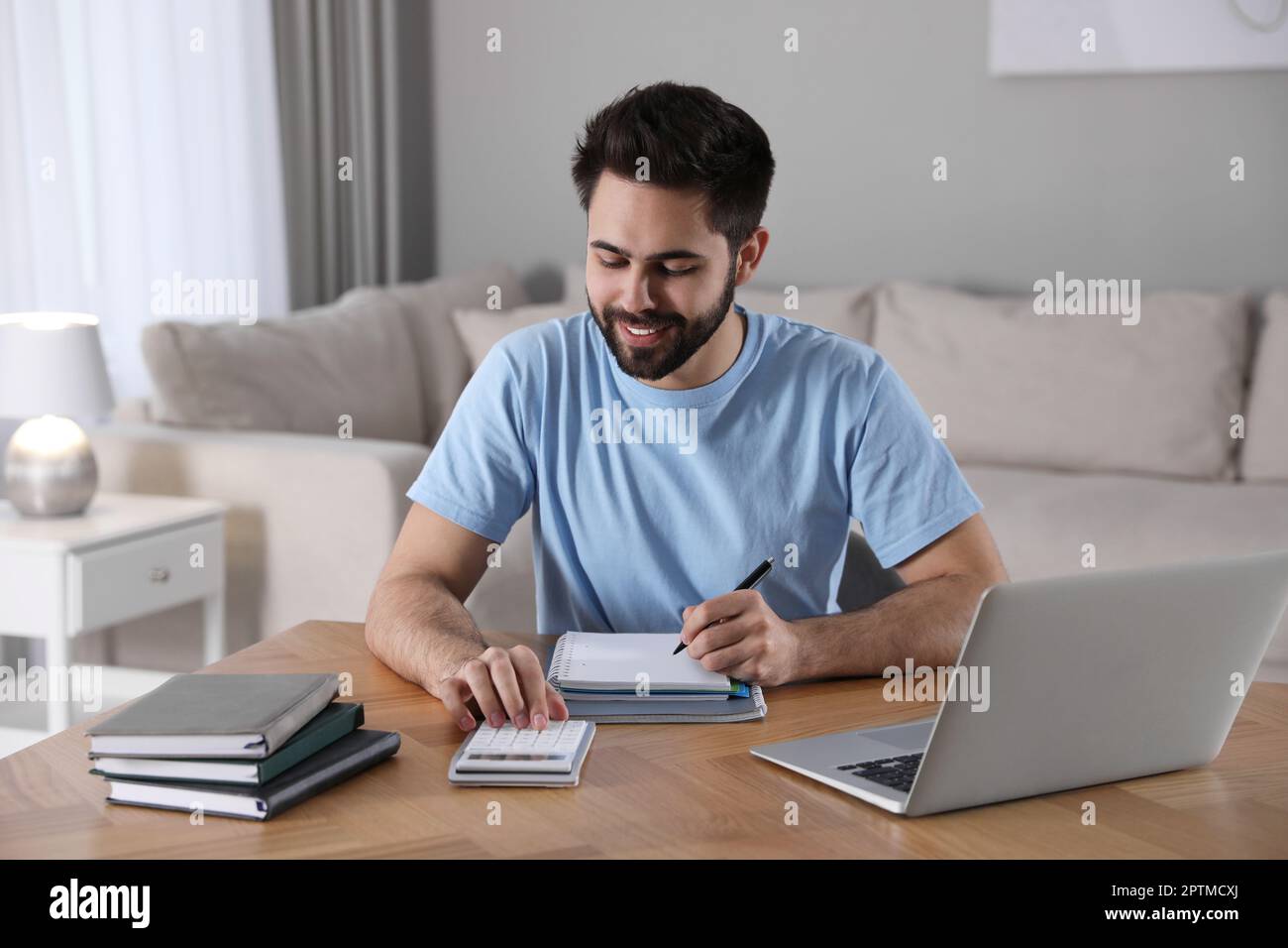 Giovane uomo che conta sulla calcolatrice durante il webinar al tavolo in camera Foto Stock