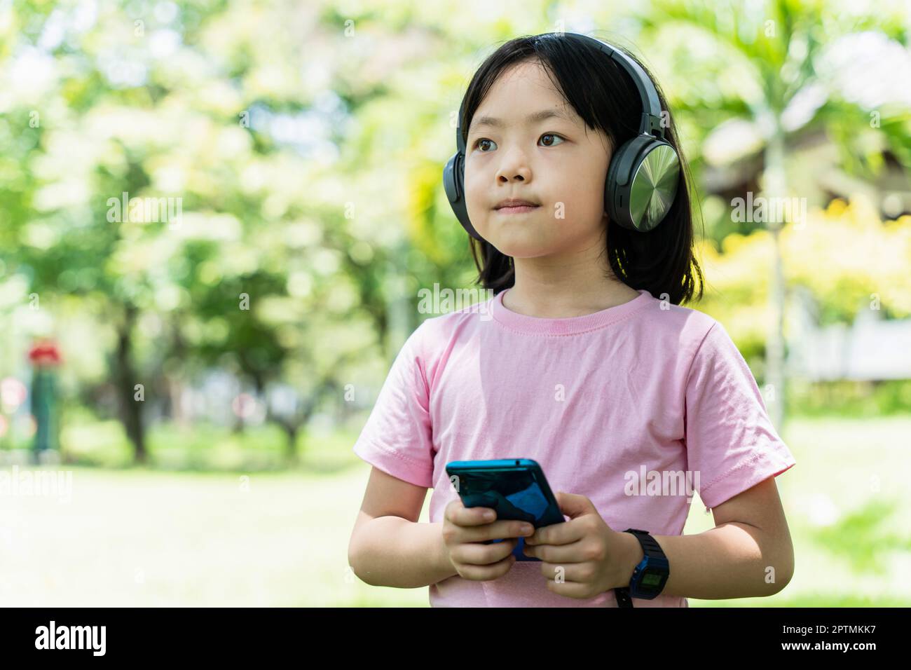 Primo piano della bambina asiatica con smartphone e ascolta la musica con le moderne cuffie wireless in un parco all'aperto. Foto Stock