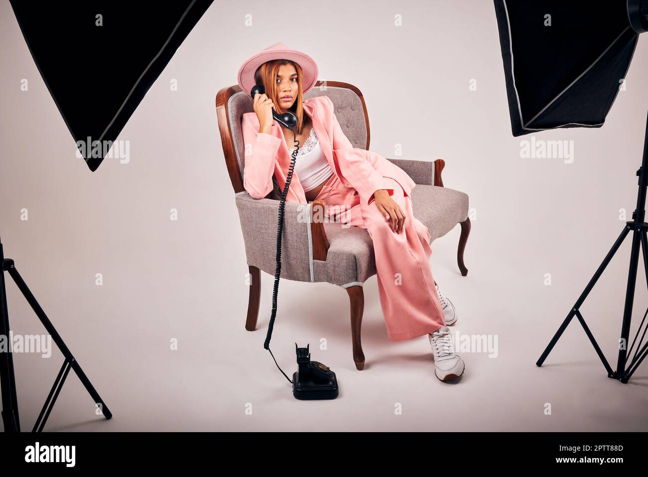 Giovane donna di razza mista elegante in posa in abiti rosa alla moda seduti su una sedia e fare una chiamata con un telefono retrò per uno studio di fotografia. FEM Foto Stock