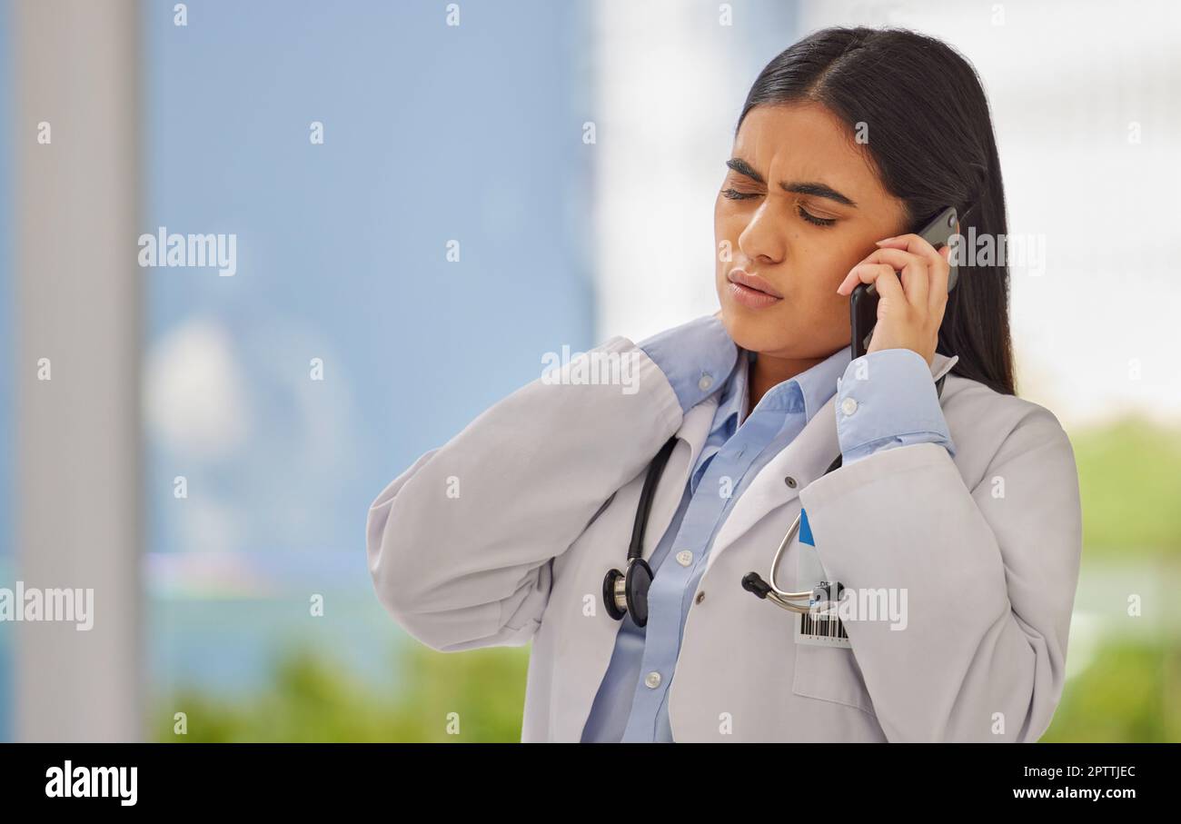 Medico stressato che parla sul cellulare in una clinica. Operatore sanitario indiano in piedi da solo e che soffre di dolore al collo in un turno occupato. Medico Foto Stock