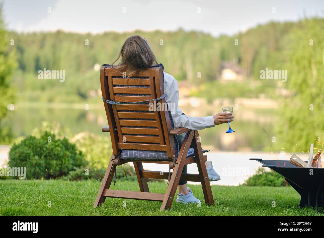 Giovane donna con un bicchiere di limonata in mano seduta accanto a un barbecue sulla riva del fiume, vista posteriore Foto Stock
