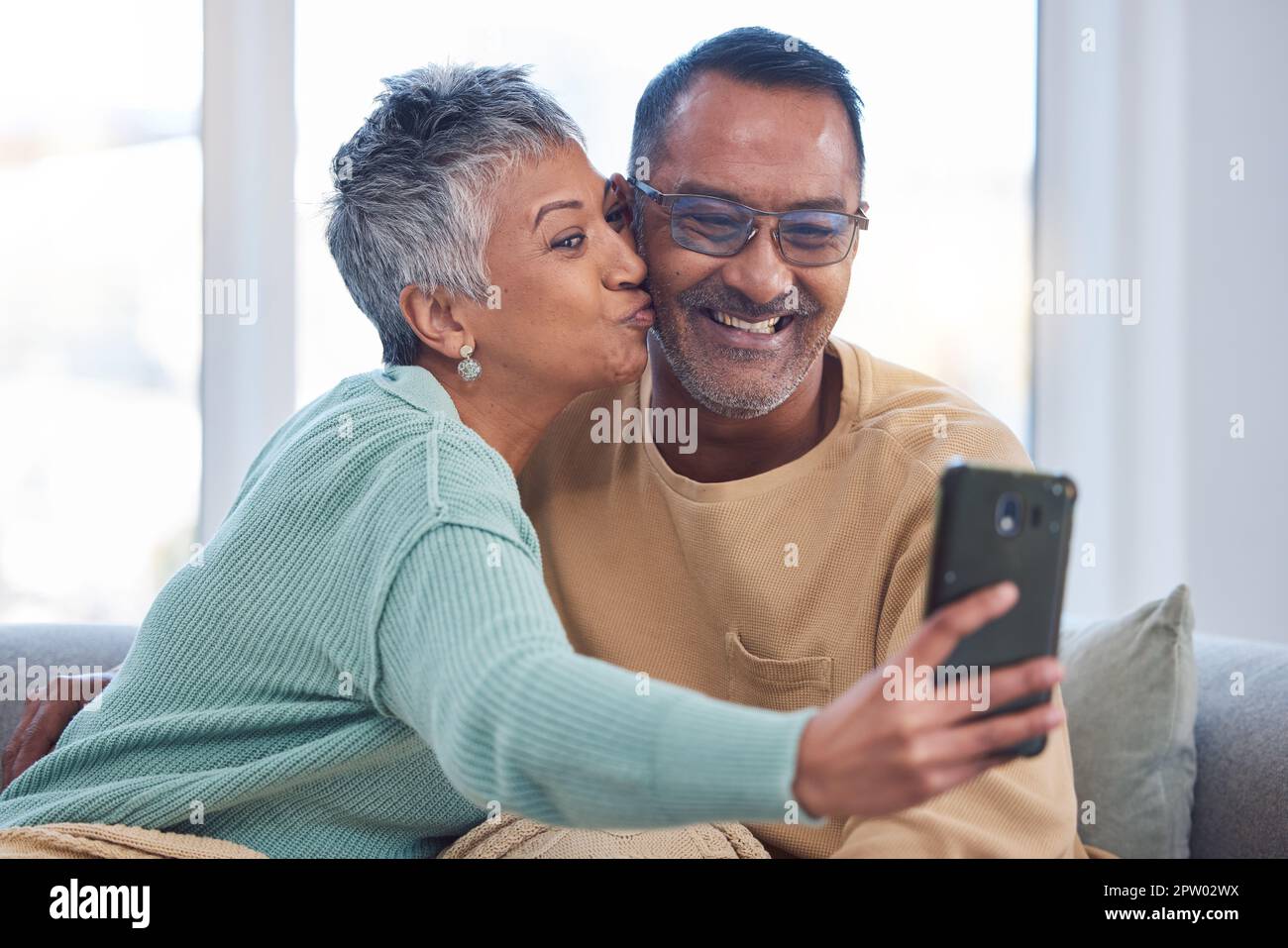 Telefono, selfie e bacio con una coppia anziana in posa per una foto nel soggiorno della loro casa insieme. Smartphone, baci e social media con Foto Stock