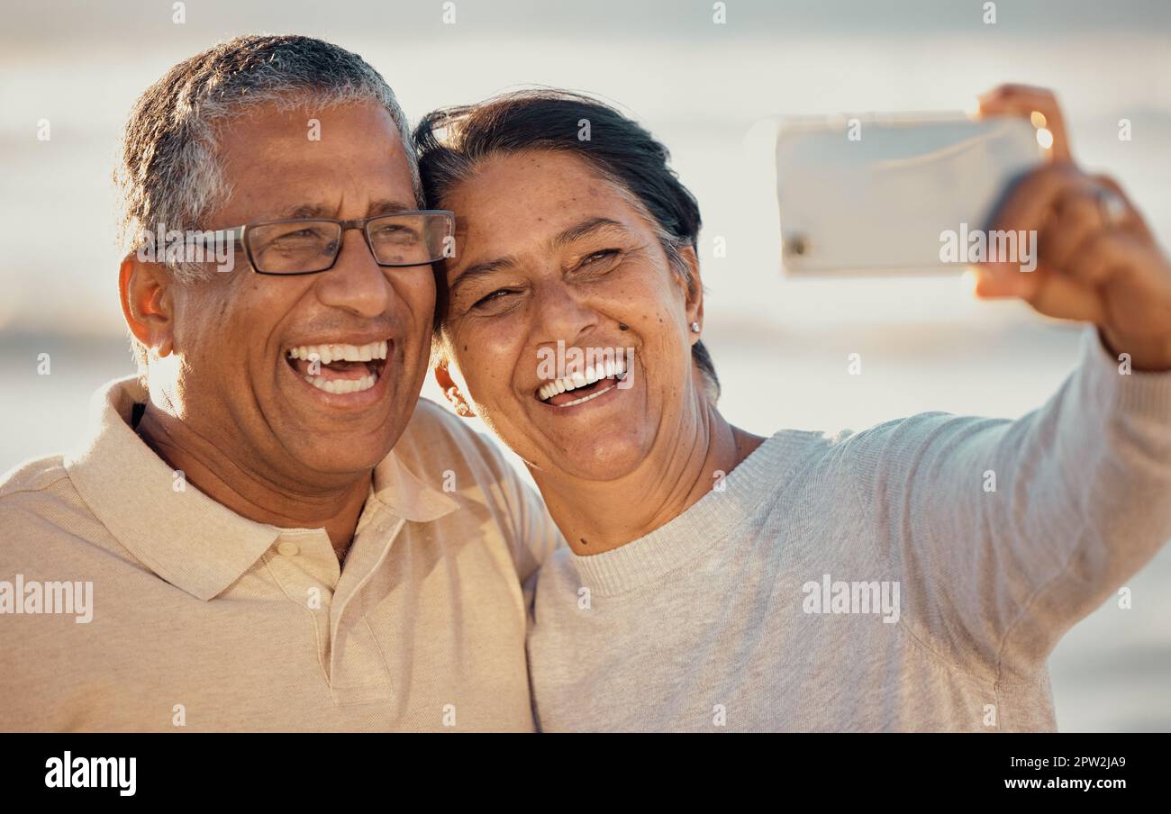 Primo piano di una coppia di corse miste affettuose senior in piedi sulla spiaggia e sorridendo e prendendo un selfie con uno smartphone durante il tramonto all'aperto. Ciao Foto Stock