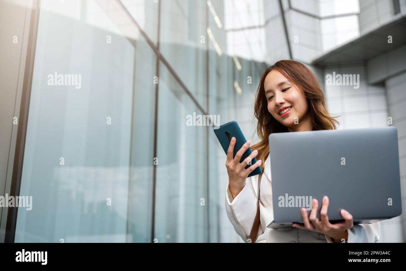 Donna d'affari sicura che utilizza smartphone con computer portatile in piedi in città. Bella donna d'affari pronta a lavorare. Concetto aziendale. Foto Stock