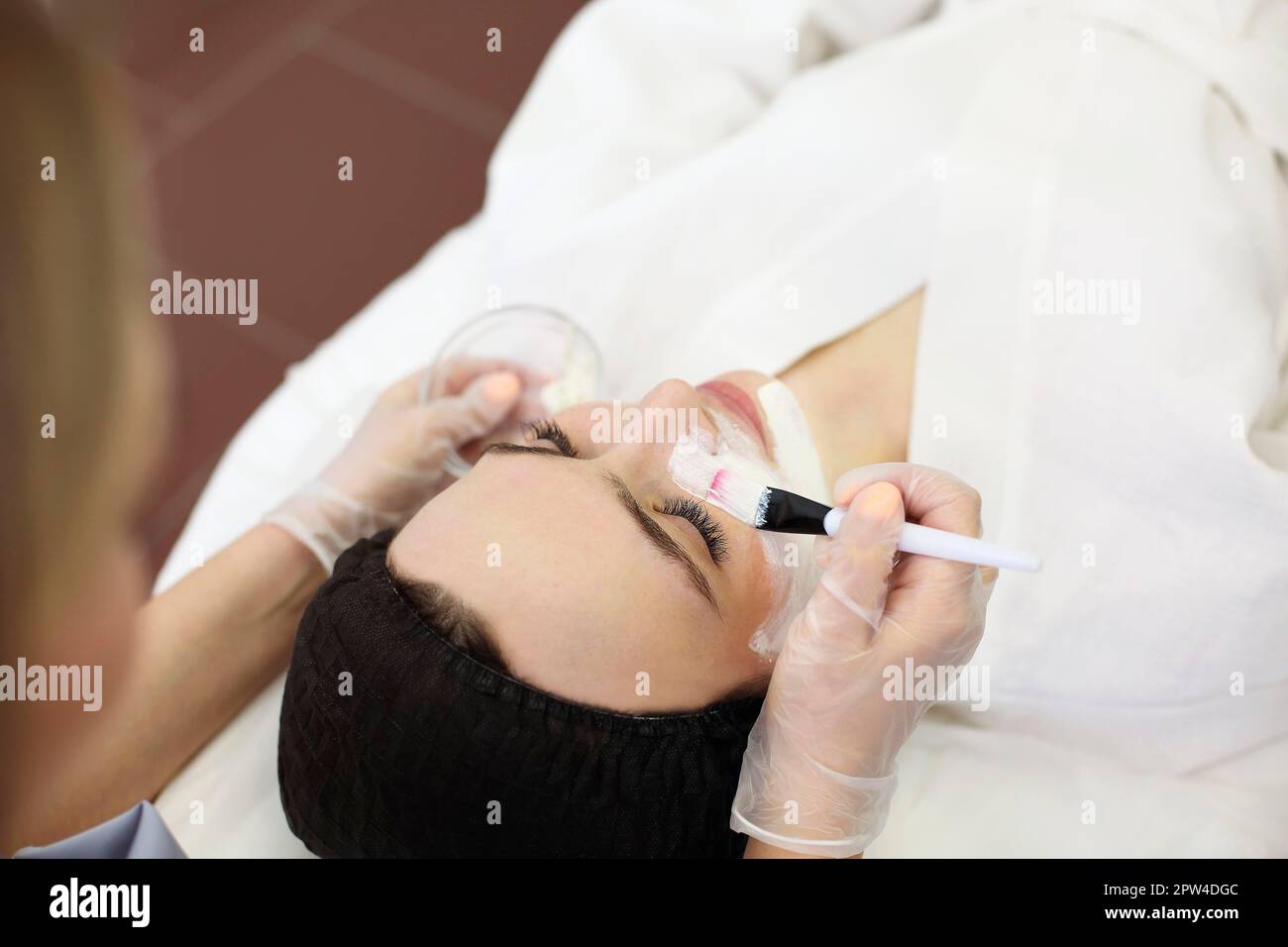 Ragazza inriconoscibile estetista che applica maschera di argilla con pennello sul viso di cliente rilassato mentre facendo la procedura di cura della pelle nel salone di cure estetiche Foto Stock