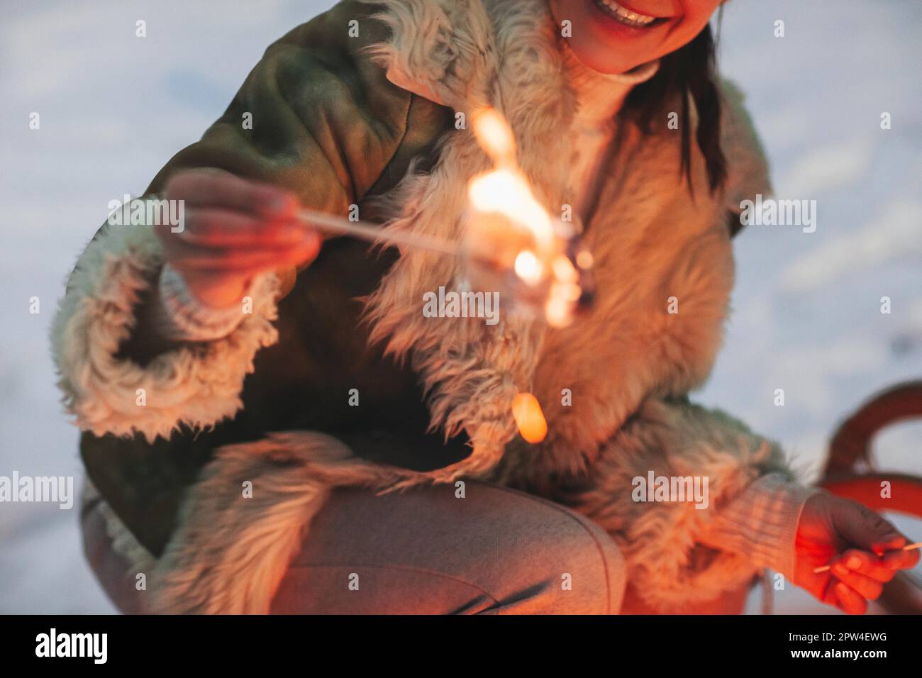 Colpo corto di donna felice arrostendo frittura dolce marshmallow sopra il fuoco del campo nella foresta di inverno, primo piano su femmina sorridente che prepara il cibo mentre Foto Stock