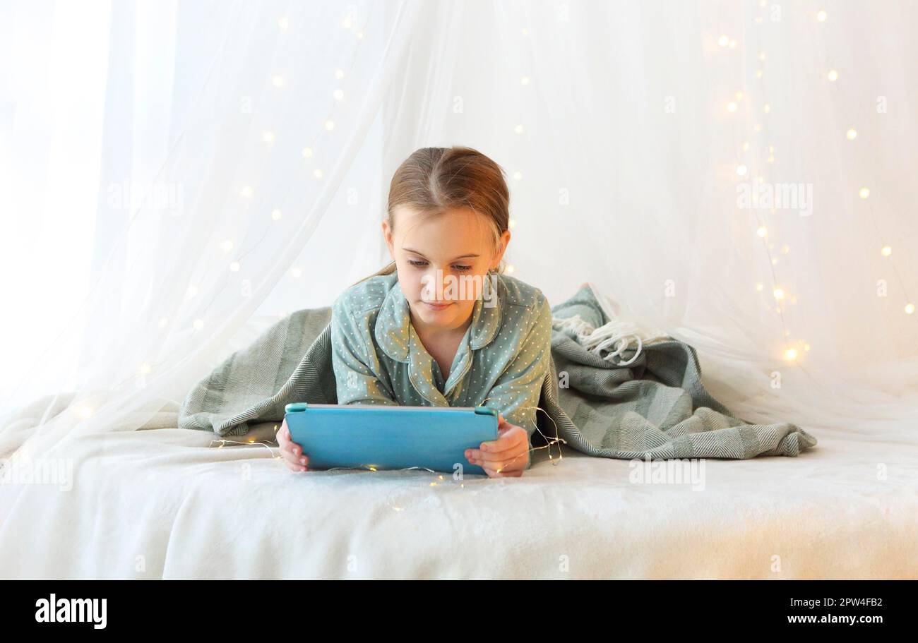 Bambini e tecnologie moderne. Bambina sdraiata in pajama con moderna tavoletta digitale su letto a baldacchino principessa decorata con luci gialle di Natale Foto Stock