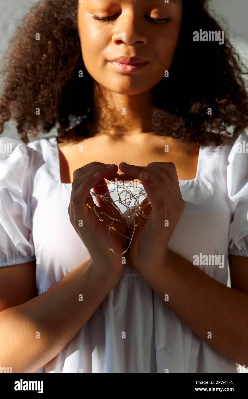 Giovane pacifica riconoscente ragazza afroamericana grata con gli occhi chiusi in vestito bianco che fa segno di cuore, luci a LED in mani di nero tranquillo Foto Stock
