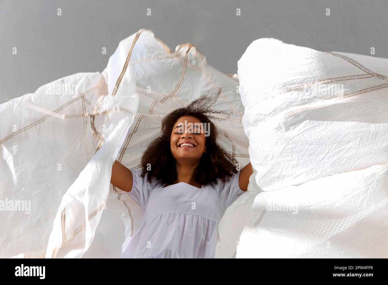 Sorridente ragazza afroamericana felice saltando con tessuto bianco in aria sullo sfondo grigio dello studio, sorridendo e ridendo ragazza nera dimostrare Foto Stock