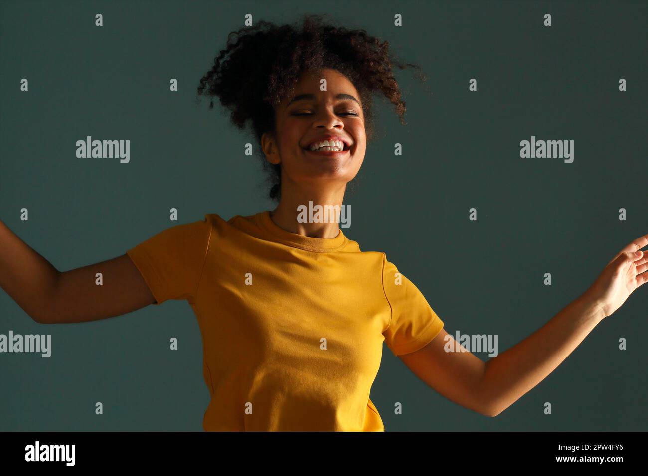 Bella donna di etnia afro-americana con pelle scura e bella in maglietta gialla che alza la mano nel saluto mentre guarda in macchina fotografica con un sorriso piacevole Foto Stock