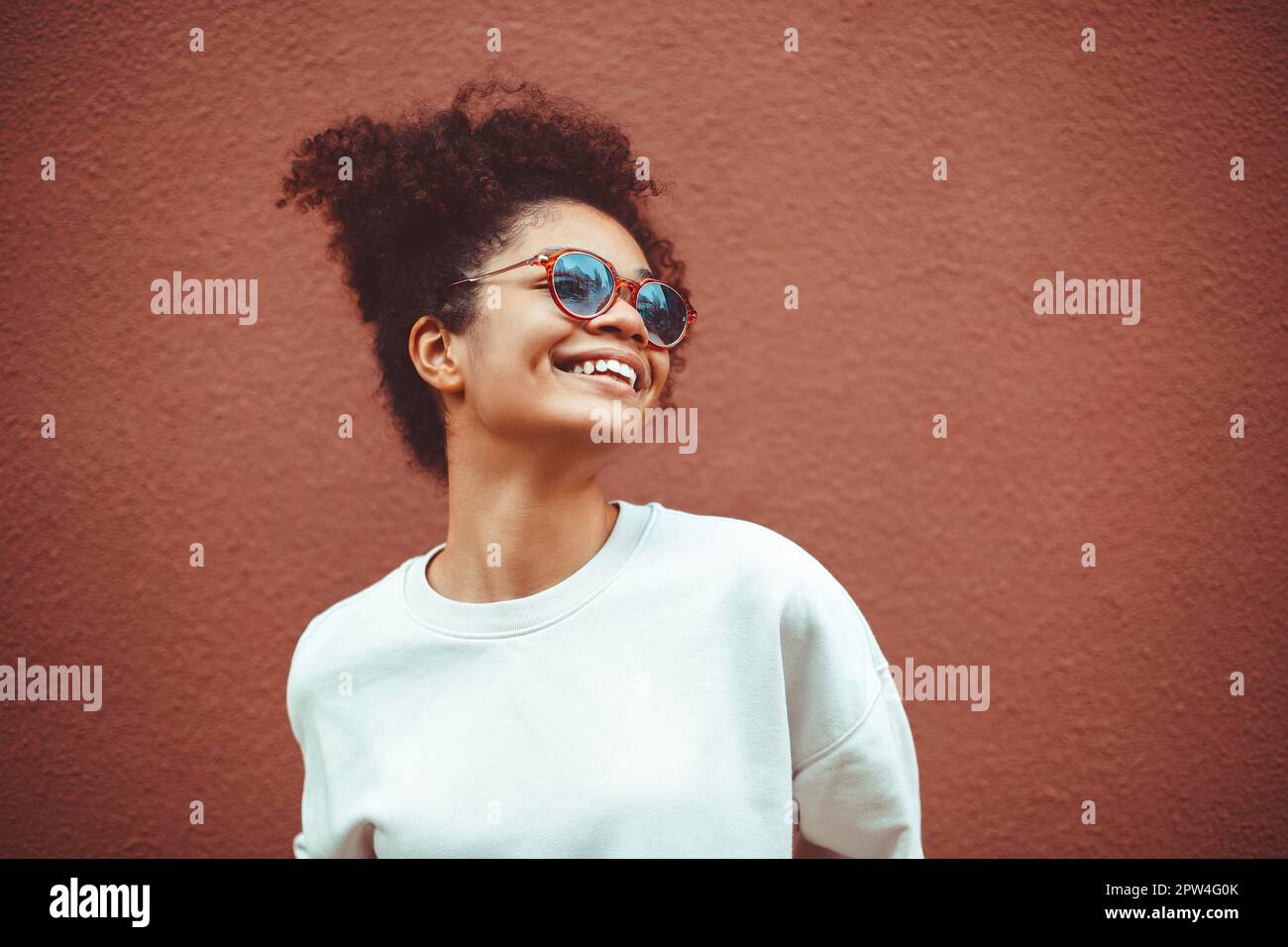 Etnia africana giovane donna in occhiali da sole alla moda, con capelli ricci legati in alto ponytail, guardando lontano mentre sorridendo ampiamente mostrando dritto Foto Stock