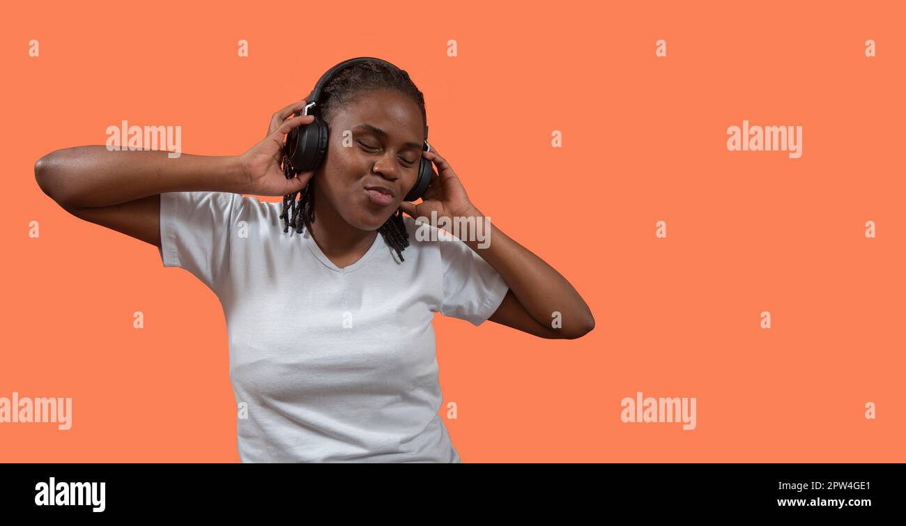 Primo piano ritratto di felice giovane donna africana che ascolta musica con le cuffie, gli occhi chiusi e tenendo le cuffie con le mani, vestito di t-shi bianco Foto Stock