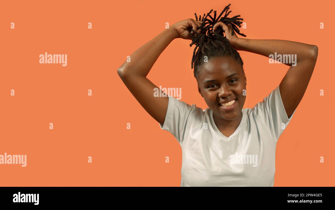 Primo piano ritratto di felice giovane donna africana guardando la macchina fotografica con grande sorriso, giocando con i capelli con le mani sollevate, vestito di t-shirt bianca e. Foto Stock