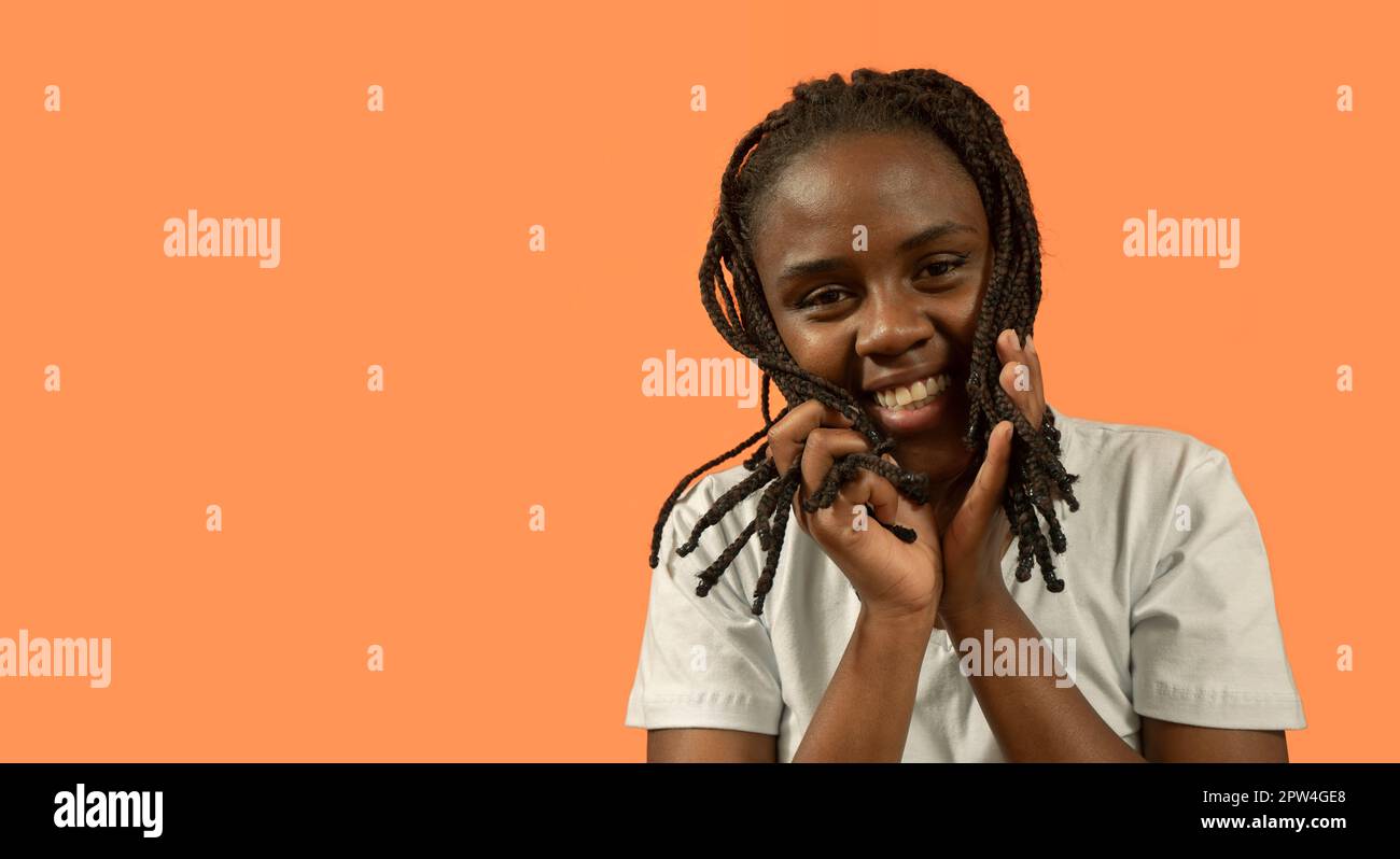 Primo piano ritratto di felice giovane donna africana guardando la macchina fotografica con grande sorriso, giocando con i capelli con le mani, vestita di t-shirt bianca e bun ha Foto Stock