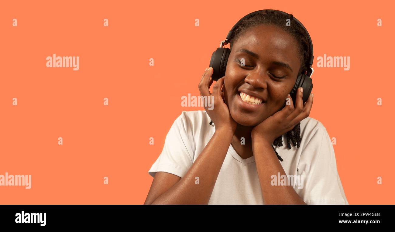 Ritratto di felice giovane donna africana che ascolta la musica su cuffie con un sorriso toothy, tenendo cuffie con le mani, vestito in t-shirt bianca agai Foto Stock