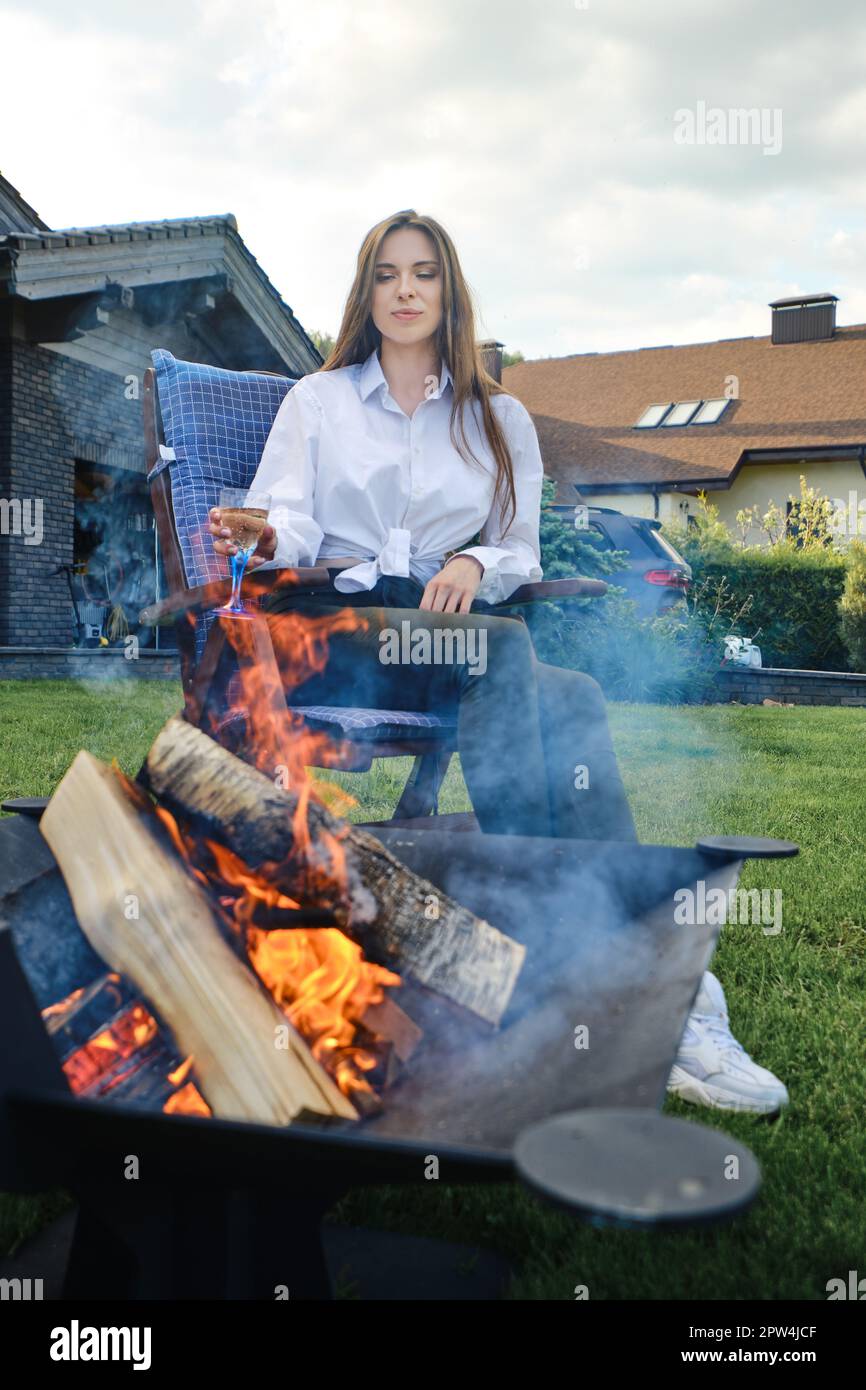 Giovane donna che guarda la legna nella griglia. È seduta in sedia pieghevole con un bicchiere di bevanda fredda in mano. Foto Stock