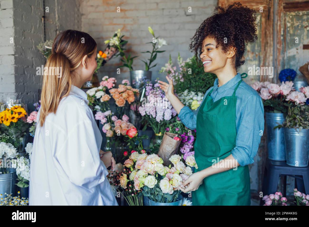 Felice imprenditore nero donna in piedi in negozio di piante che vende fiori freschi al cliente. Giovane ragazza bionda comprare un bouquet fresco da fiorista. Sorridente Foto Stock