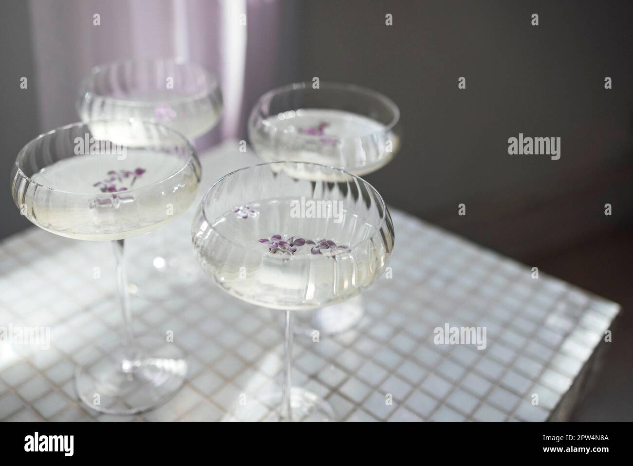 Quattro bicchieri con champagne su una superficie di un tavolo leggero che riflette la morbida pioggia del sole. Vino frizzante con fiori di lilla. Vista dall'alto Foto Stock