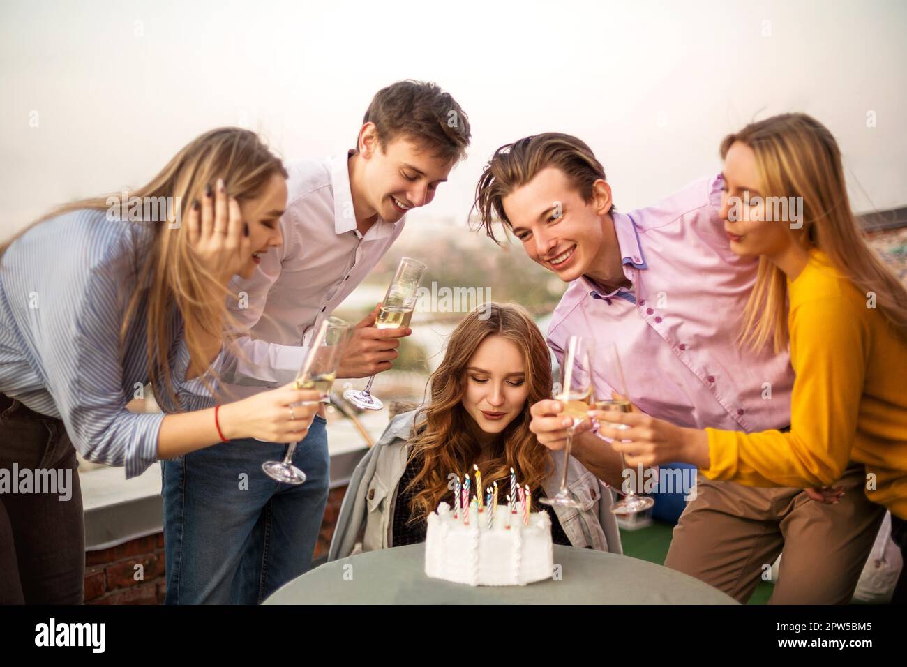 Giovane bionda felice ragazza soffia candele nel suo compleanno fare un desiderio sul tetto circondato da amici gioiosi Foto Stock