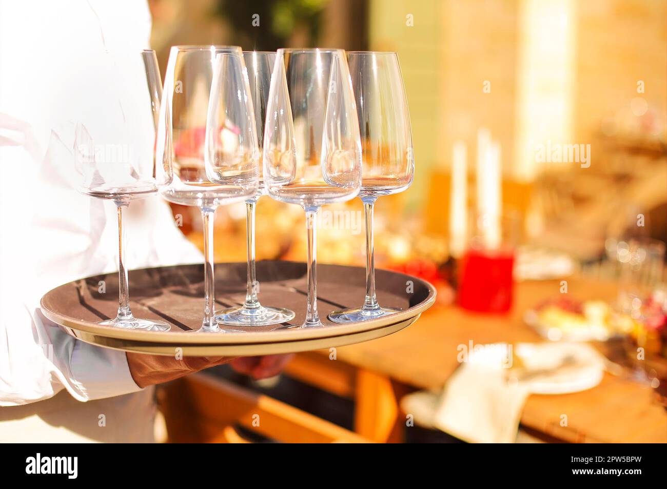 Foto ritagliata del cameriere dal servizio di catering indossando una camicia bianca che trasporta un vassoio rotondo marrone con set di bicchieri da vino vuoti puliti al tavolo dei clienti Foto Stock