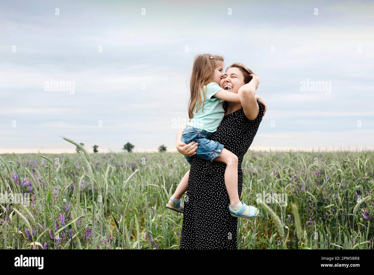 Felice madre e figlia con fiori di campo bouquet abbracciando in verde prato. Madre che abbraccia una bambina all'aperto in un campo. Amore e famiglia Foto Stock